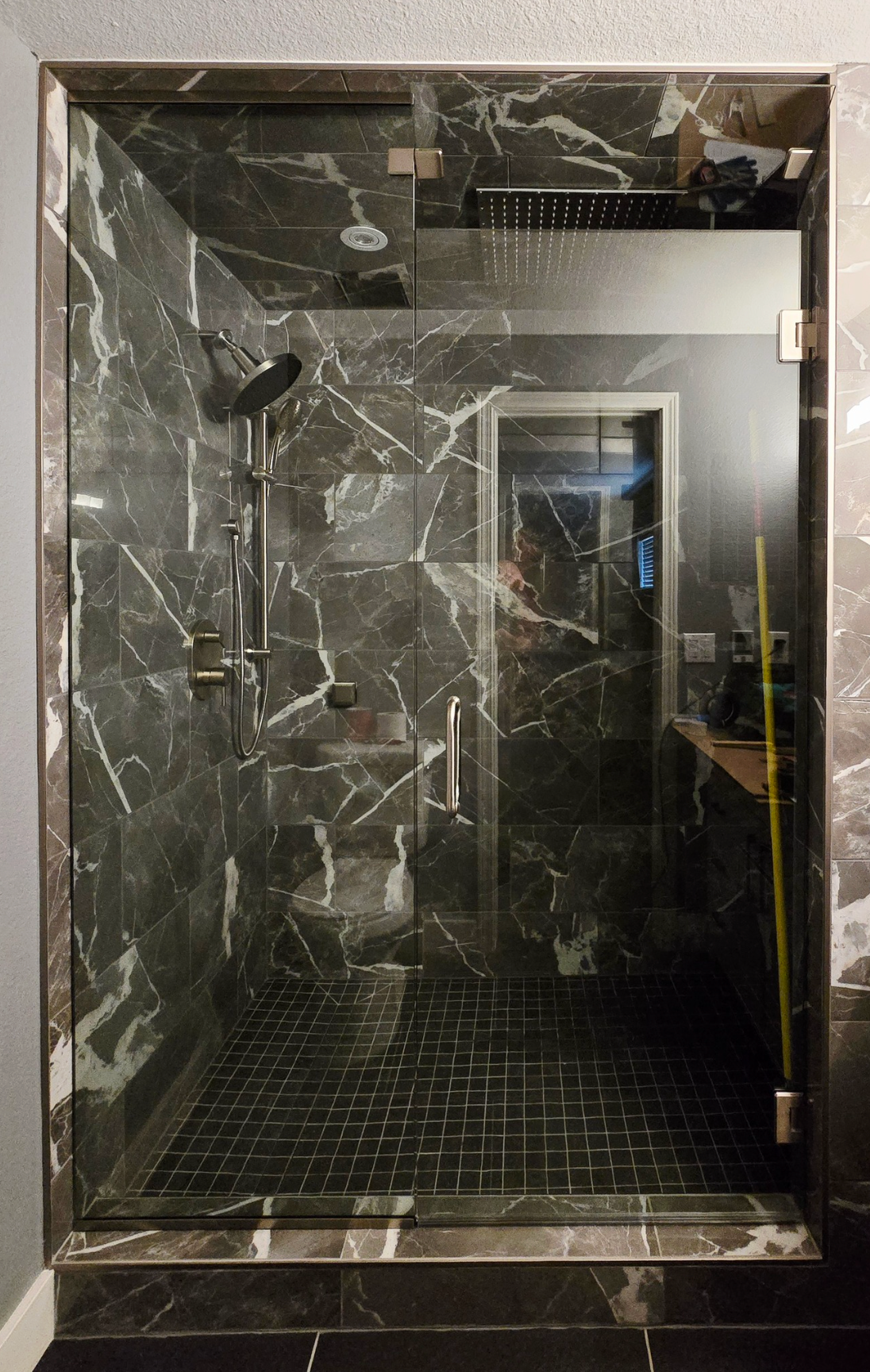 A glass-enclosed shower with dark marble walls and tile flooring, featuring a silver showerhead and a handheld sprayer.
