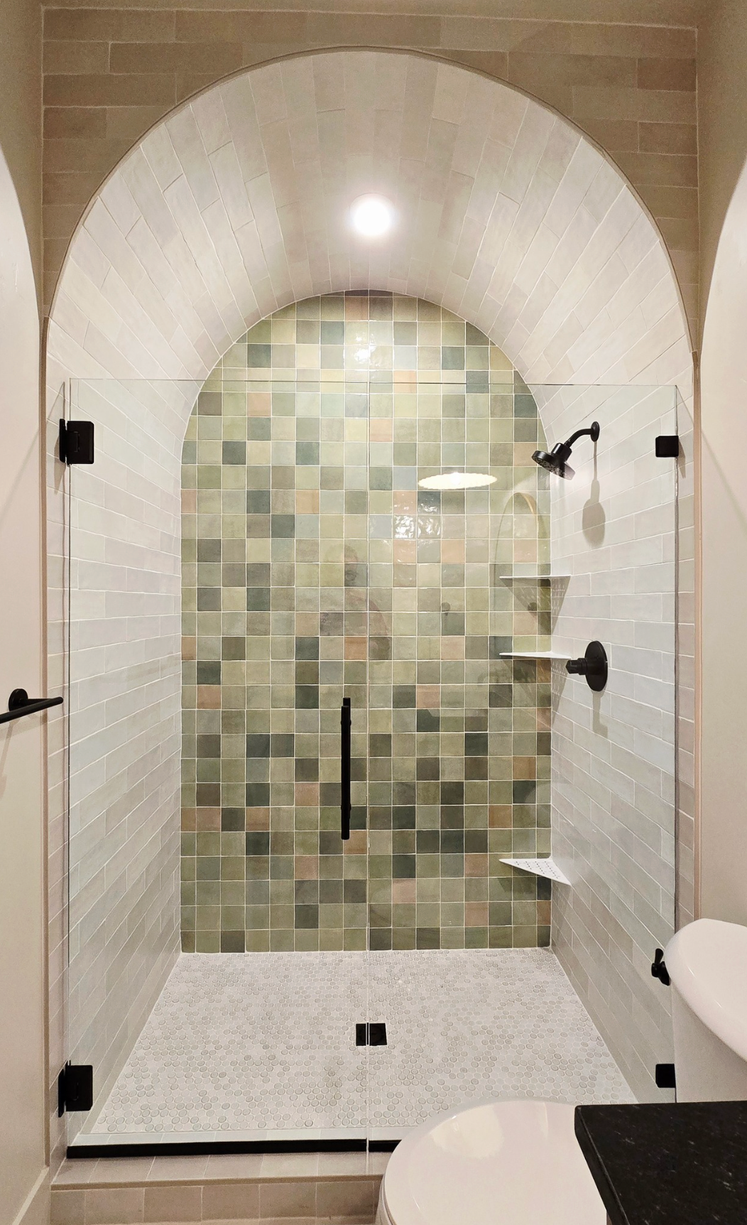 An arched walk-in shower with a mosaic tile back wall, white subway tile sides, a glass door, and black fixtures.