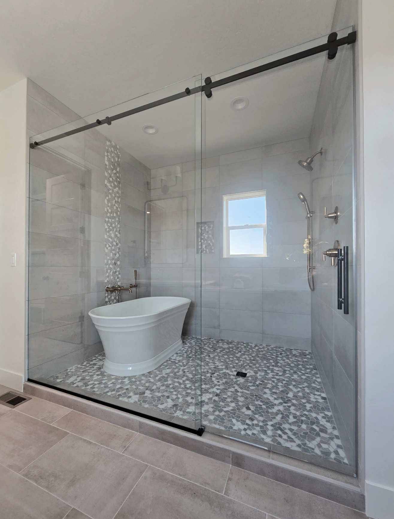 A modern walk-in shower with a freestanding white bathtub, gray tile walls, mosaic floor, and a glass sliding door.