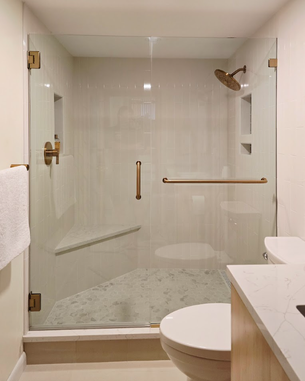 A modern glass shower enclosure with brass fixtures, a built-in corner bench, and a towel on a rack next to a toilet.