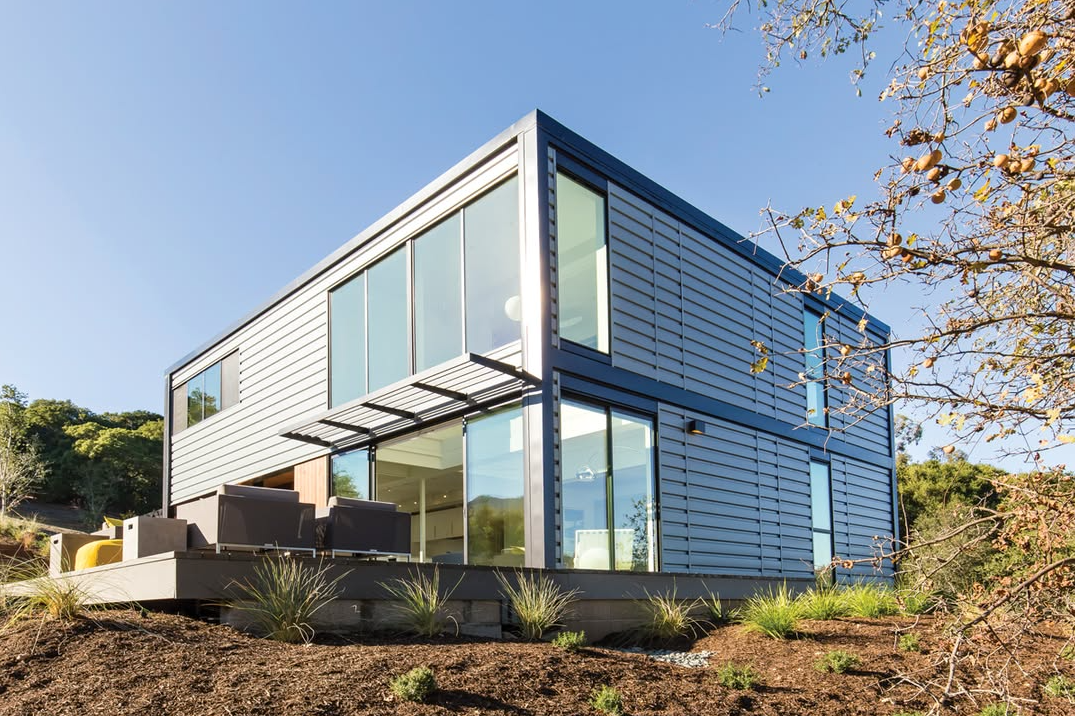 Modern two-story house with large glass windows, white siding, and a patio in a sunny hillside setting