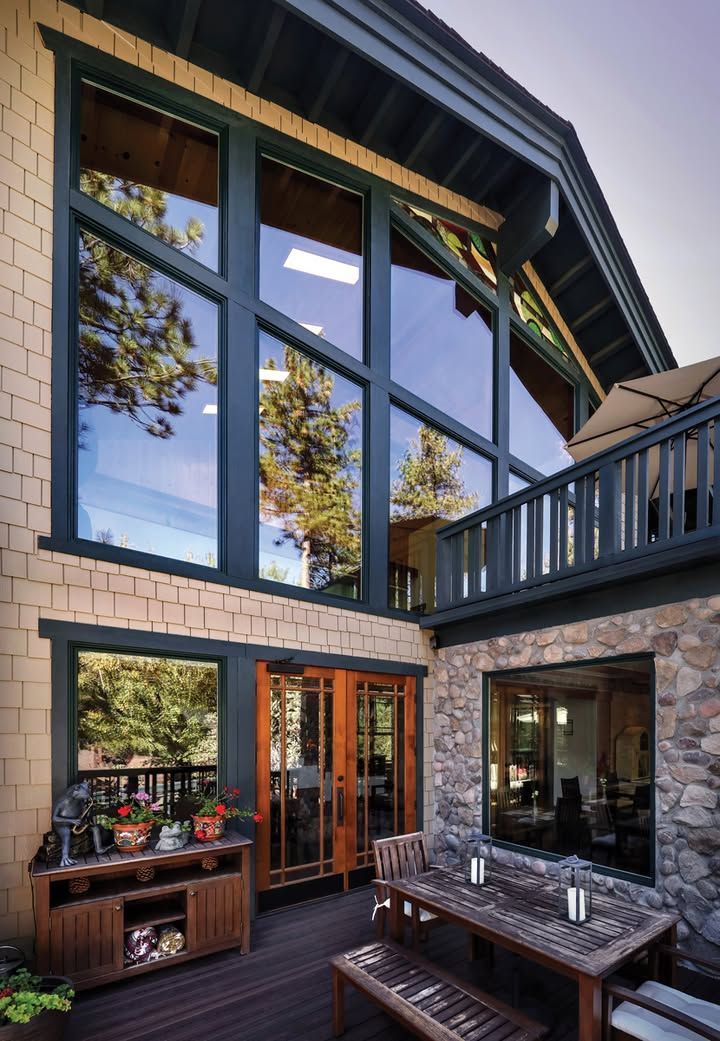 A wooden patio with a dining set and a console table beside a house with large windows, stone siding, and a balcony.
