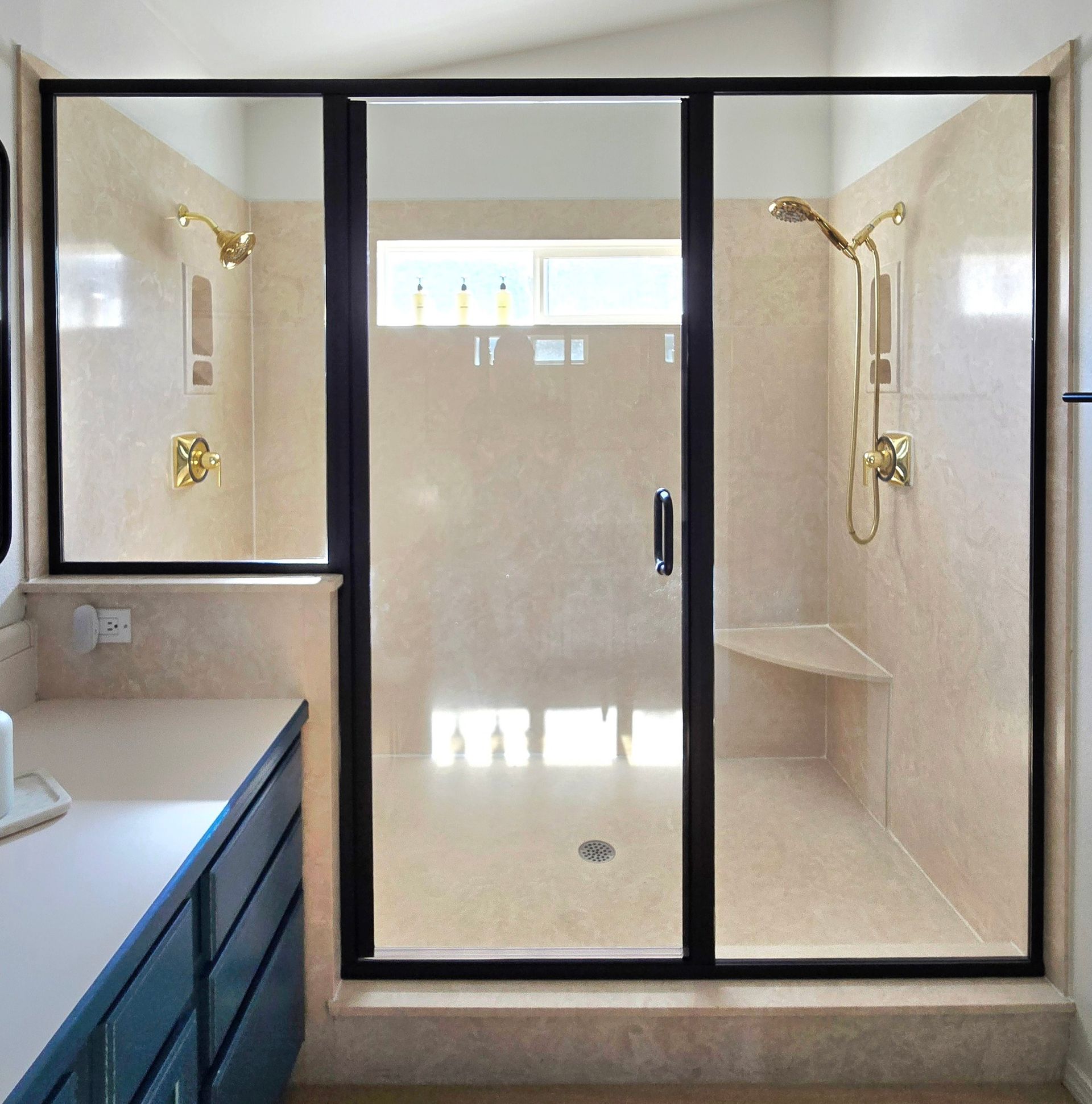 Bathroom shower with black frame, gold fixtures, beige tile, and a built-in seat.