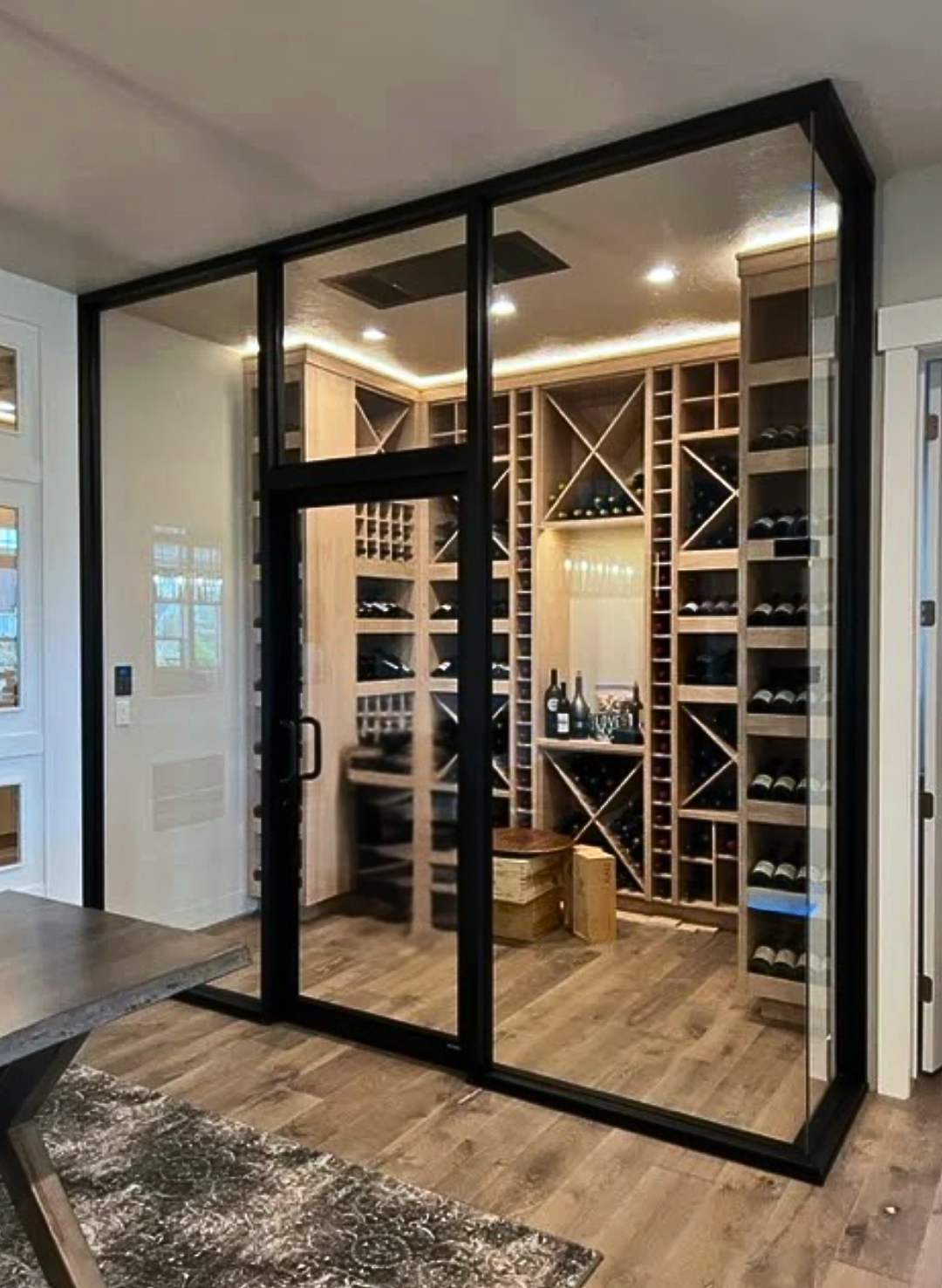 Glass-enclosed wine cellar with wooden racking and bottles, black frame, interior view.