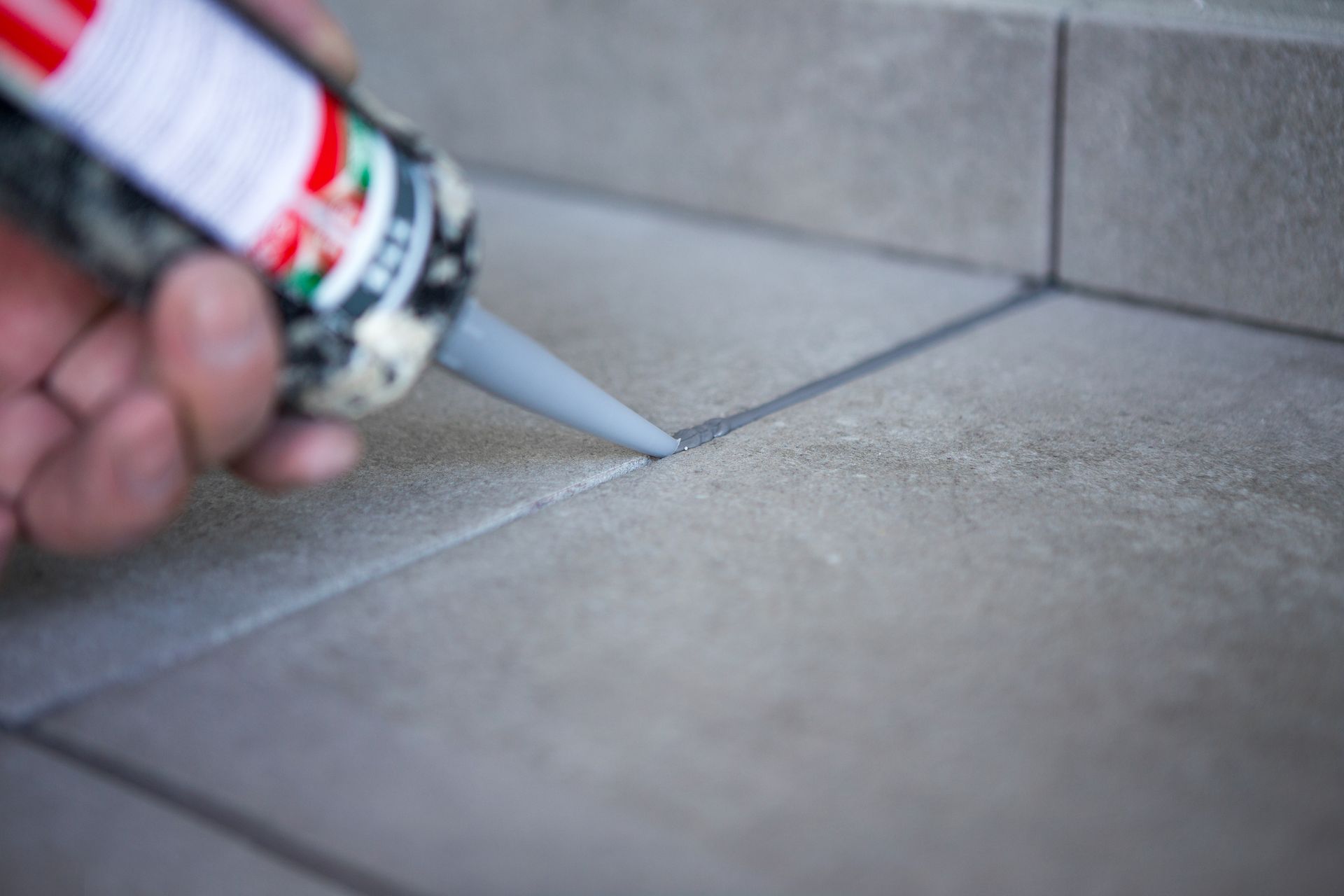 Hand applying caulk to grout lines between gray floor tiles. Hand applying caulk to grout lines between gray floor tiles.