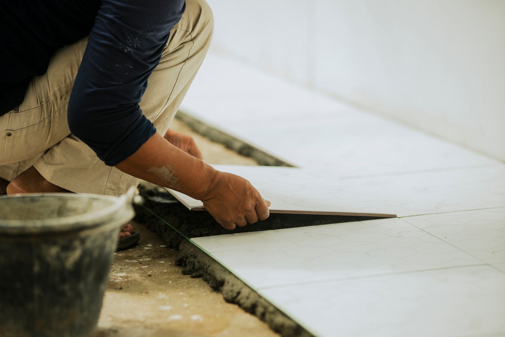 Person laying white tiles on a floor, with a bucket in the foreground. Person laying white tiles on a floor, with a bucket in the foreground.