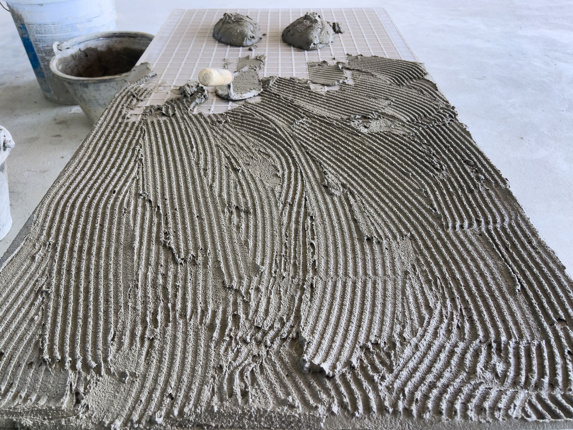 A rectangular tile surface covered in grooved mortar, with globs of mortar and a bucket nearby. A rectangular tile surface covered in grooved mortar, with globs of mortar and a bucket nearby.