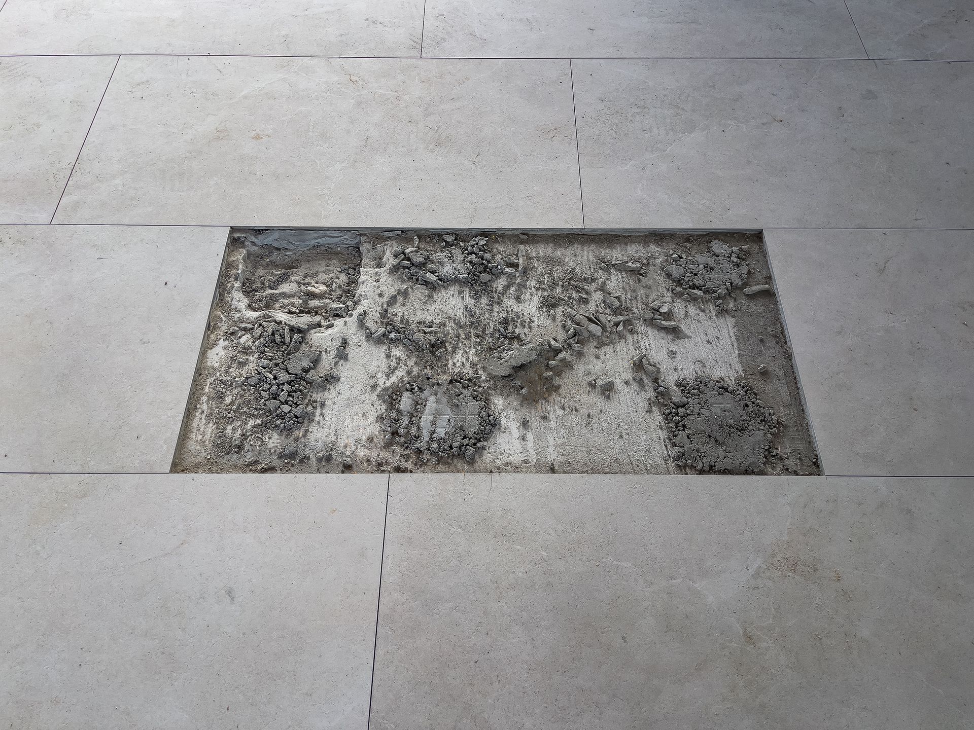 A rectangular inset in light-colored stone paving is filled with rough, gray material. A rectangular inset in light-colored stone paving is filled with rough, gray material.
