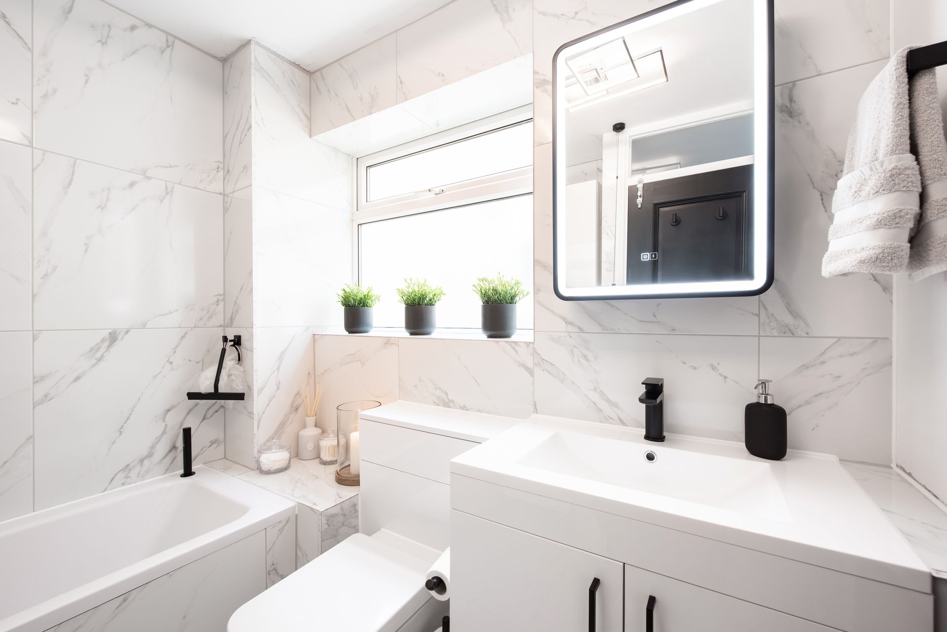 Bright white bathroom with marble-like tiles, a bathtub, toilet, vanity, and a window with plants. Black fixtures.