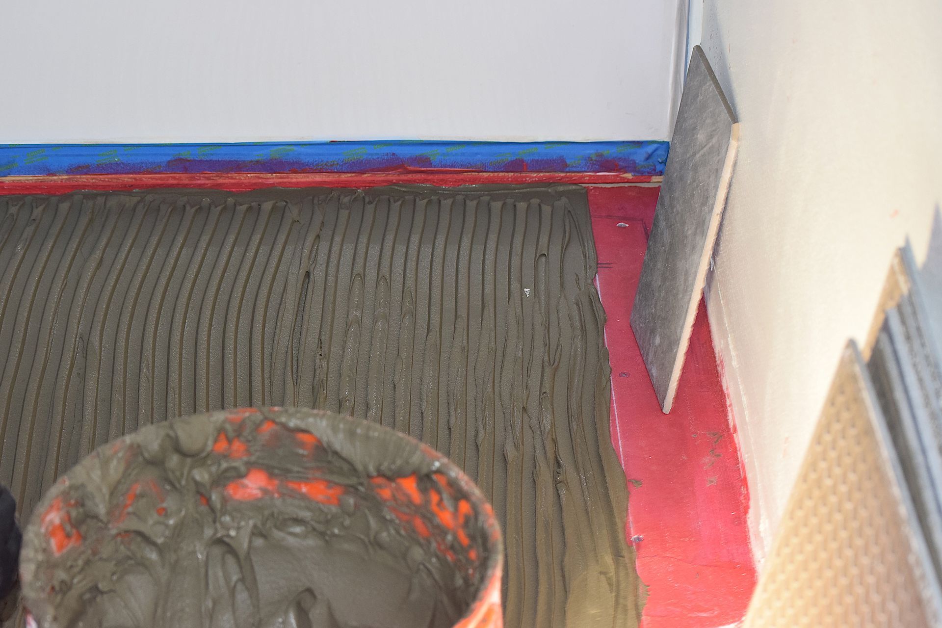 Close-up of floor with troweled mortar, bucket of mortar, and tiles being installed on a wall. Close-up of floor with troweled mortar, bucket of mortar, and tiles being installed on a wall.