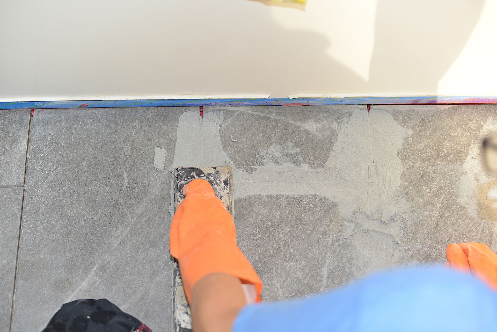 Person wearing orange gloves using a trowel to apply grout to stone tiles on a floor. Person wearing orange gloves using a trowel to apply grout to stone tiles on a floor.