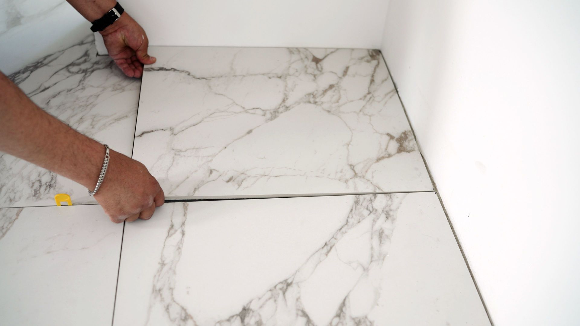 Hands placing marble-patterned tile on a floor during installation; adjacent to a wall. Hands placing marble-patterned tile on a floor during installation; adjacent to a wall.