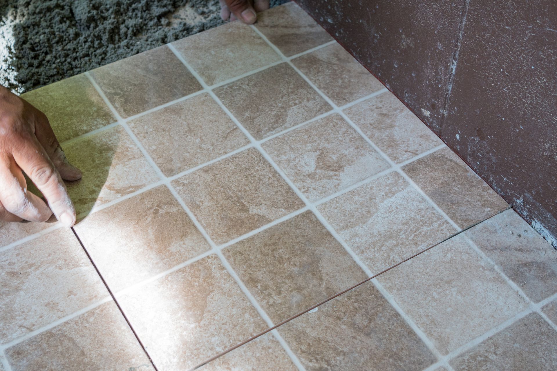 Person's hands aligning floor tiles, in a corner, with light brown square tiles.