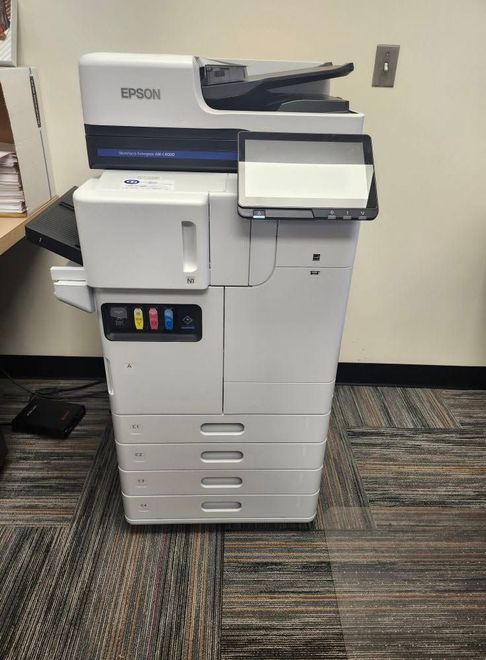 White Epson office copier/printer beside a wall in a carpeted workspace.