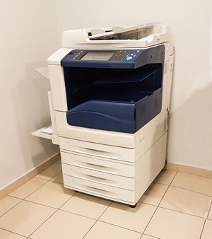 Large white office copier in a corner on a tiled floor, with paper stacked on top