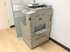Gray office copier/printer beside a wall in a hallway with light wood flooring