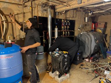 Two people working in a cluttered basement workshop with tools, drums, and equipment.