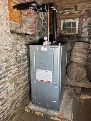 Gray furnace in a rough stone basement with exposed pipes and stacked firewood nearby