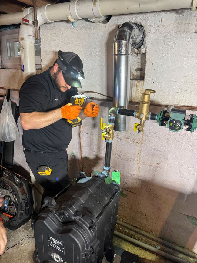 Technician using a drill on basement pipes beside a water heater and furnace