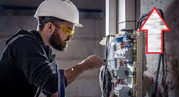 Electrician in hard hat and safety glasses working on a circuit board, wires visible on wall.