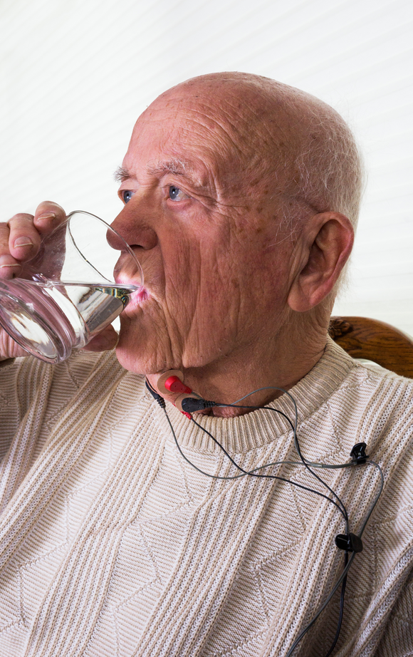 An elderly man is drinking water from a glass while wearing headphones.