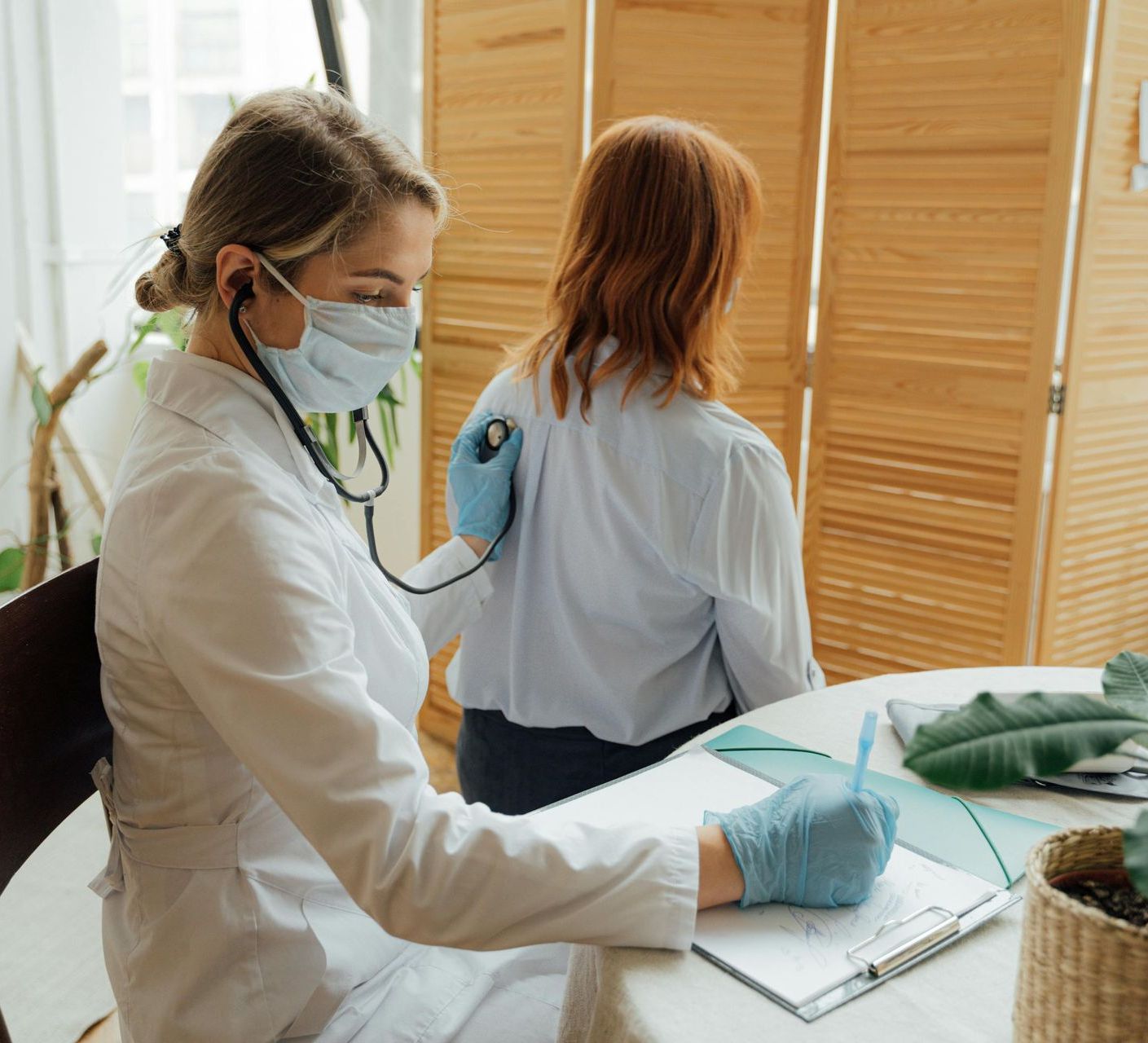 A doctor is wearing a mask and using a stethoscope to listen to a patient 's back.