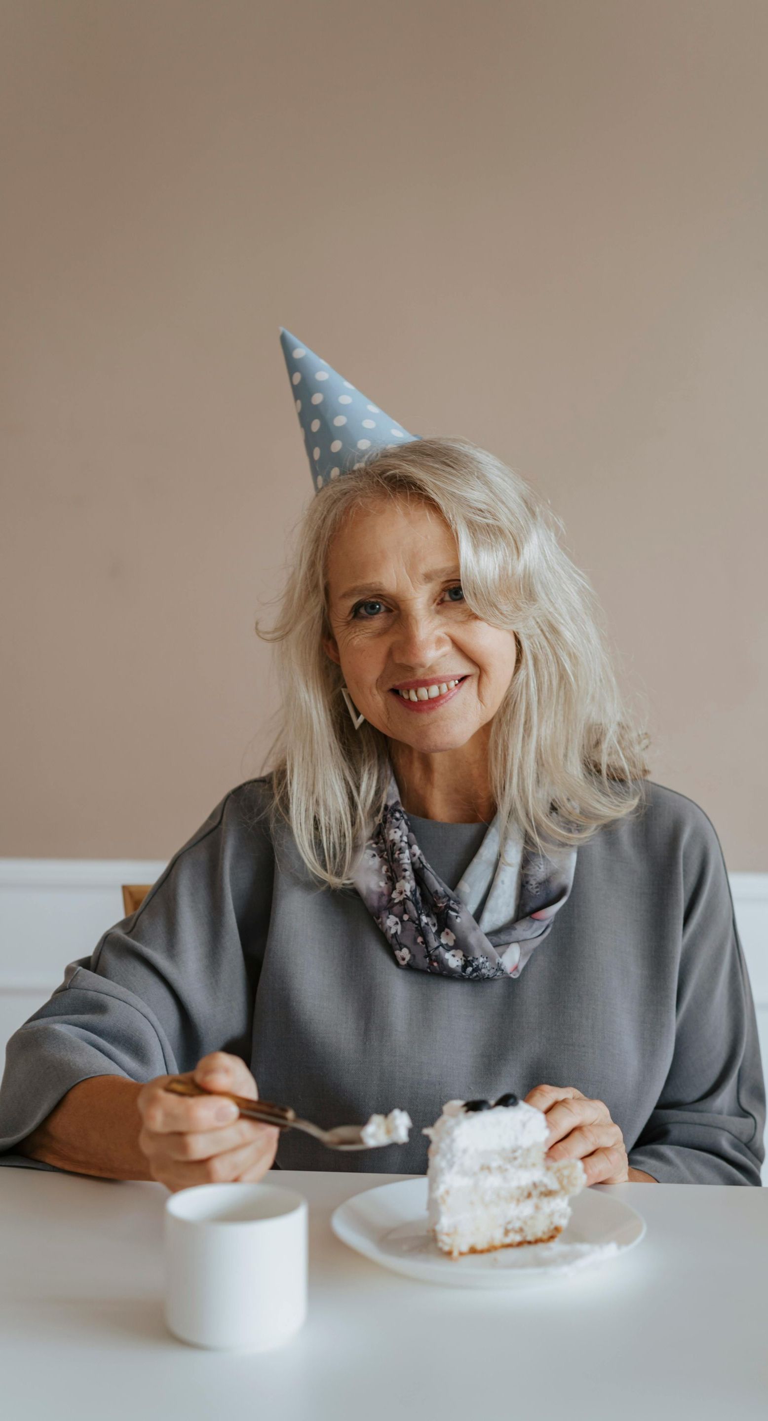 An elderly woman wearing a party hat is sitting at a table eating a piece of cake.
