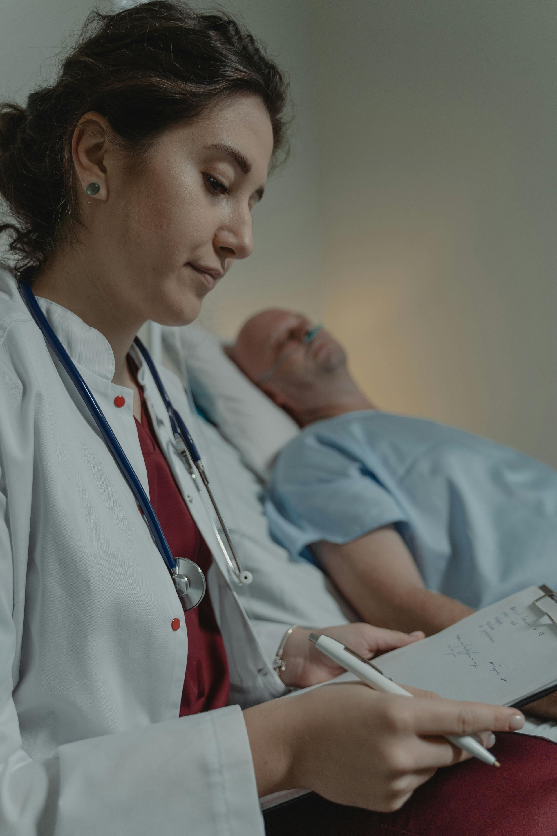 A doctor is writing on a clipboard next to a patient in a hospital bed.