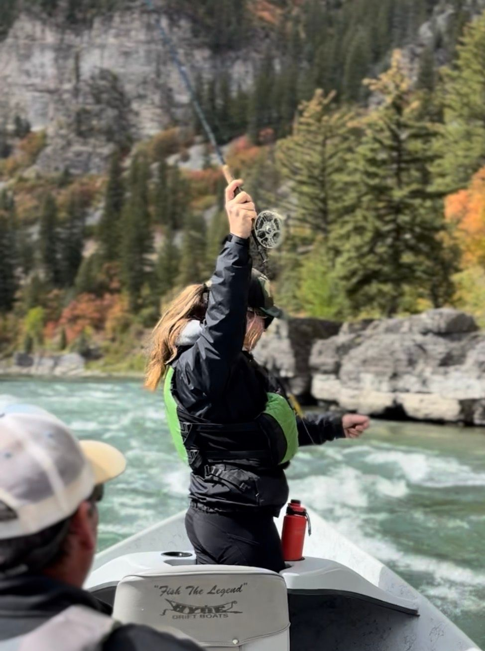 Person kayaking on a turquoise river, raising one arm, with forested cliffs in the background