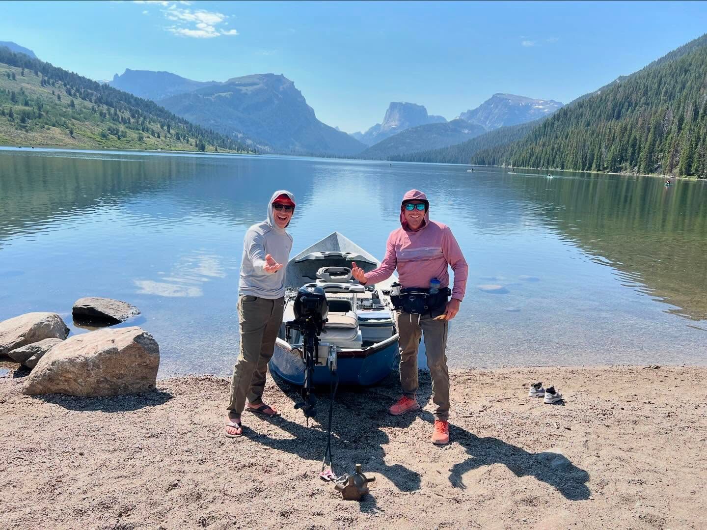 Two people pose beside a jet ski at a rocky lake shore with mountains and calm water behind them