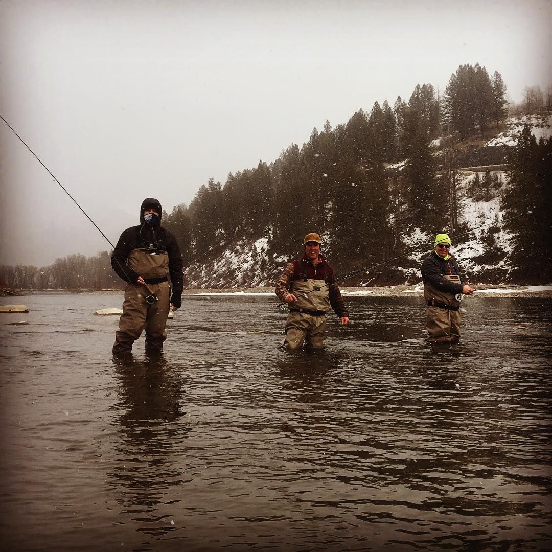 Three anglers wade in a shallow river near a forested, snowy bank, fishing in cold weather.