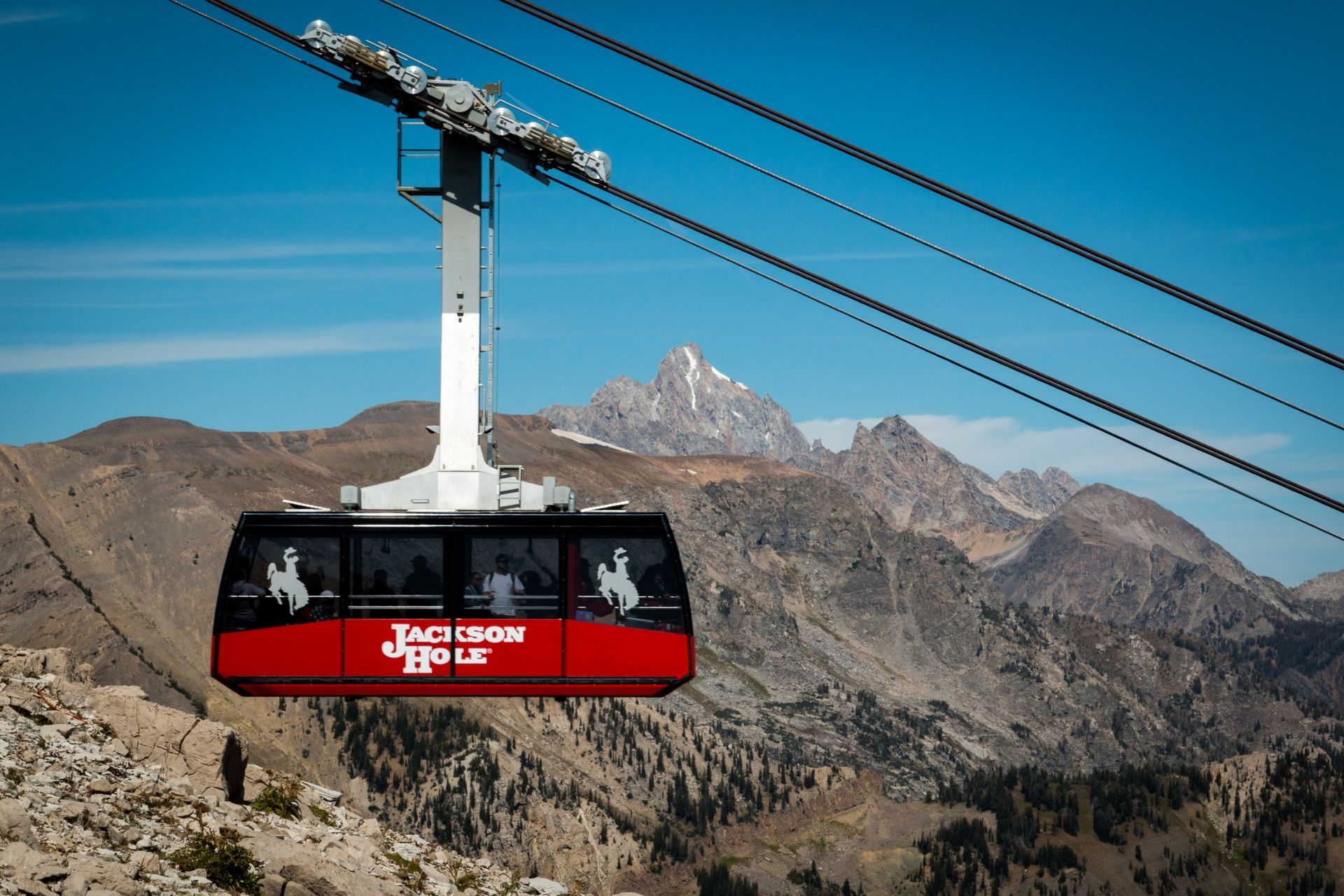 A red cable car glides above a rocky mountain landscape with snowy peaks in the background.