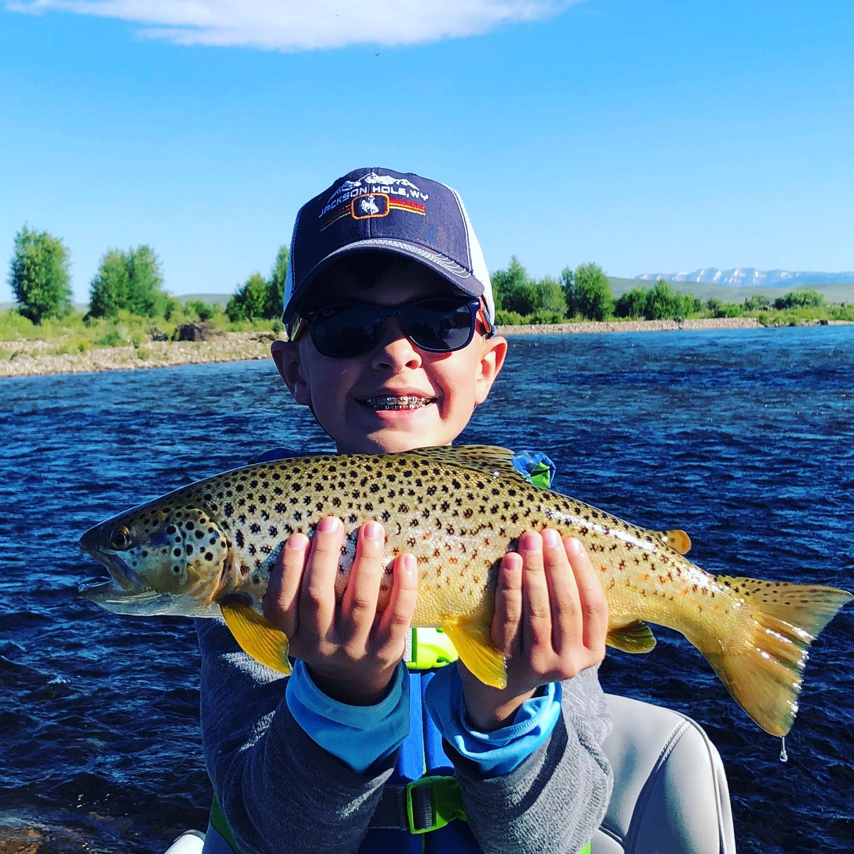 Person holding a large spotted fish in a river under a clear blue sky.