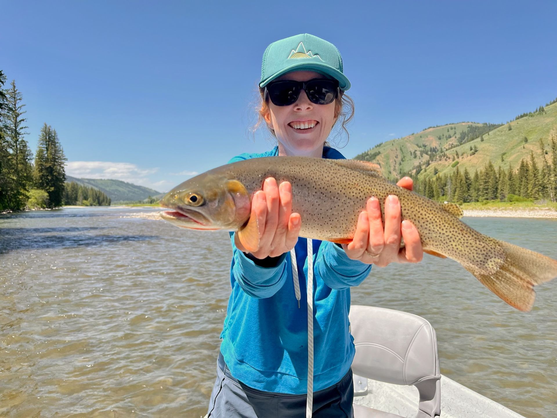 Person holding a large fish in a river, wearing a blue shirt and cap with mountains in the background