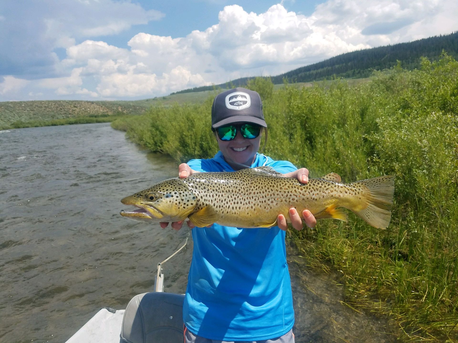 Person in blue shirt and cap holding a large fish beside a river under a cloudy sky.