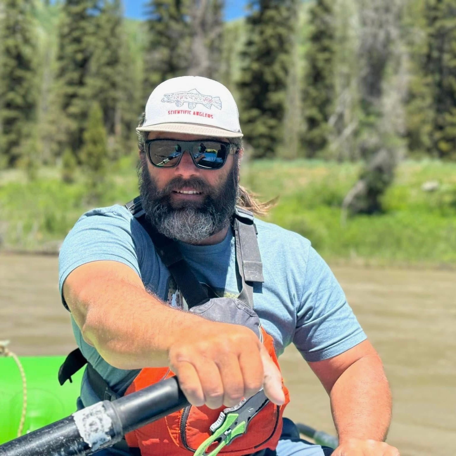 Bearded kayaker in helmet and sunglasses paddling on a green river with forest in the background