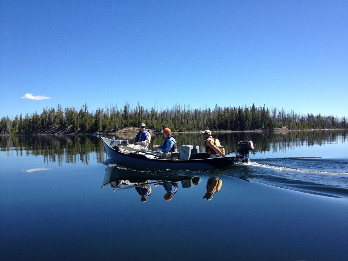 Three people in a motorboat on a calm lake, leaving a wake beside a forested shoreline under a blue sky.