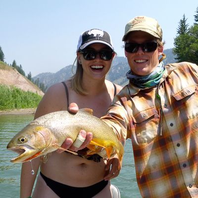 Two people smiling and holding a large fish by a river in a mountain setting