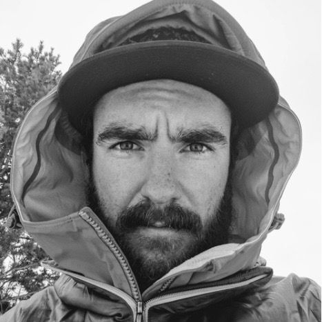 Close-up black-and-white portrait of a bearded person in a hooded jacket and cap, looking at the camera.