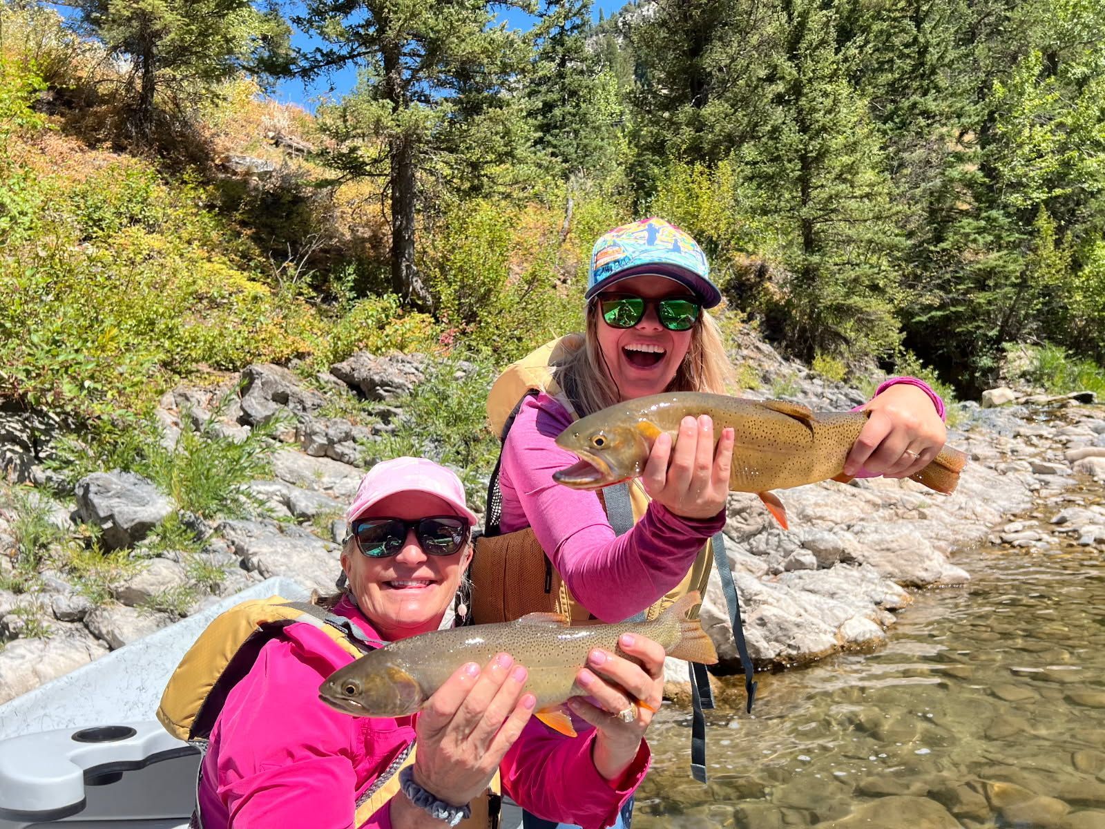 Two people in bright pink tops smiling and holding fish by a rocky stream in a forest