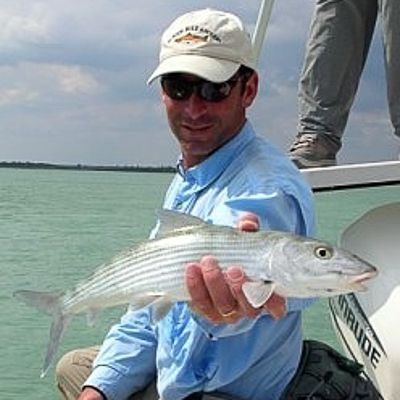 Man on a boat holds up a silver fish over green water under a clear sky