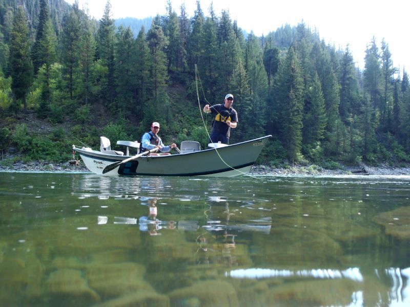 Two people fishing from a small boat on a calm river with forested shoreline