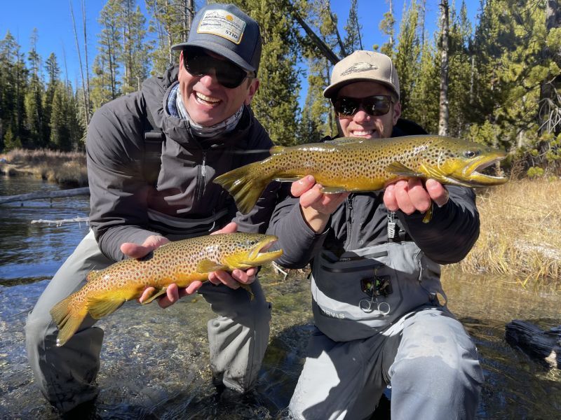 Two anglers kneel by a river, smiling and holding large golden-brown fish in a pine forest.