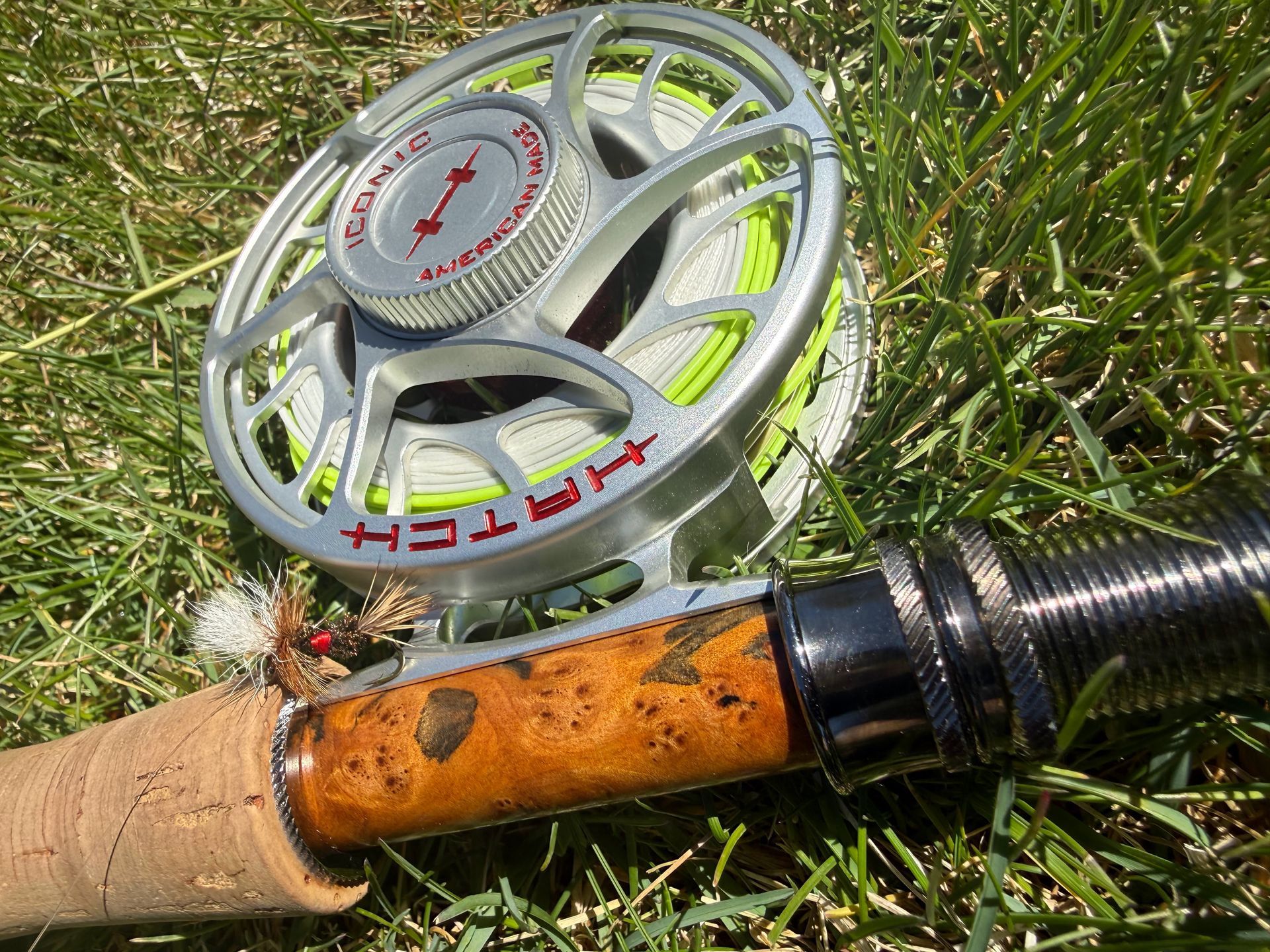 Fishing reel on a cork rod handle resting in grass, with silver and orange details.