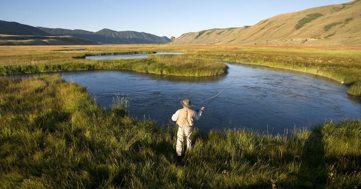 Person standing by a river in a grassy valley with hills in the background