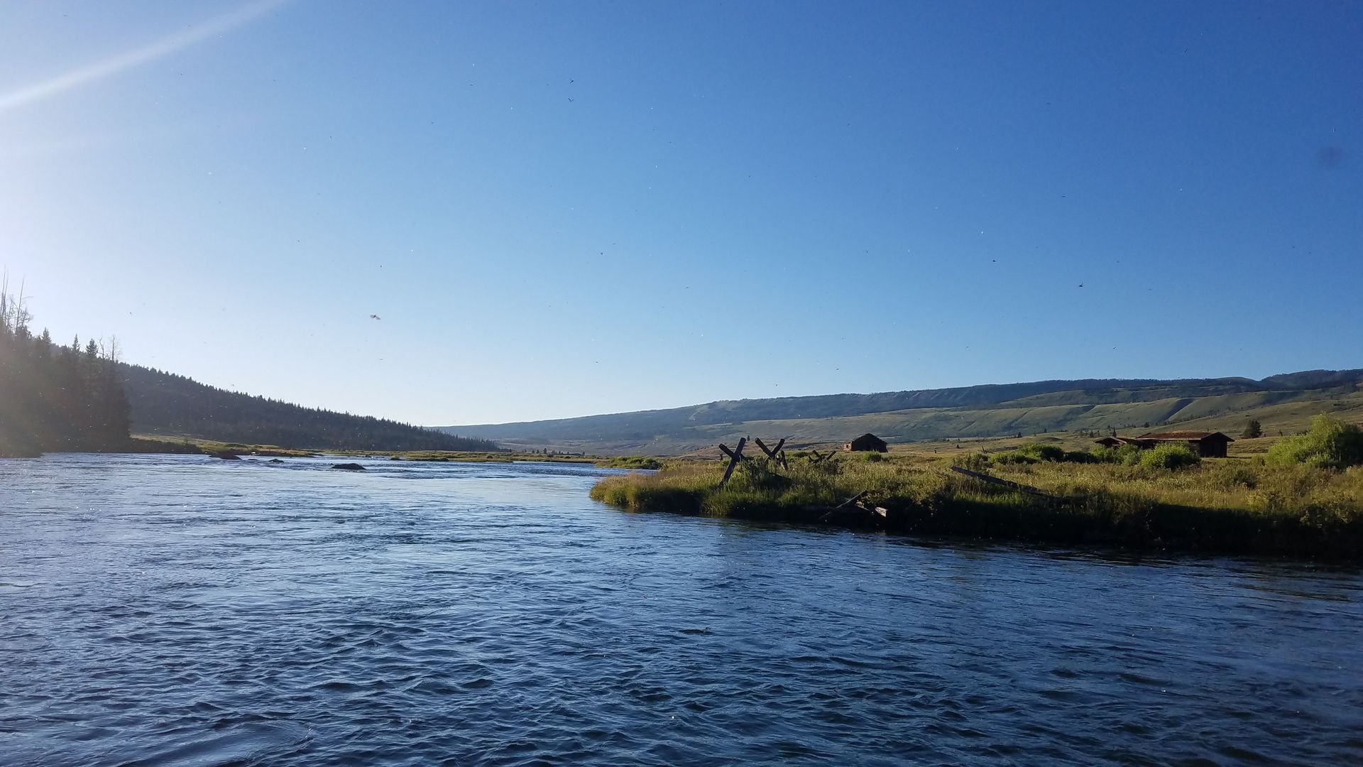 Wide river under a clear blue sky, with grassy banks and distant hills in sunlight.