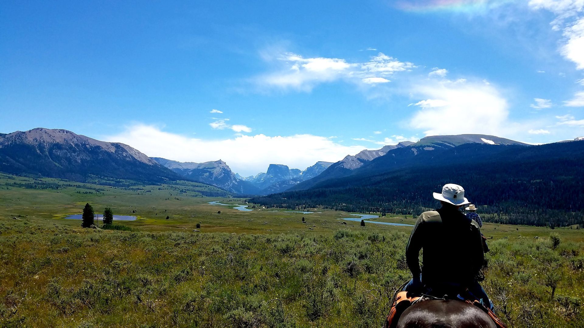 Rider on horseback in a grassy mountain valley under a blue sky, with distant peaks and a few hikers