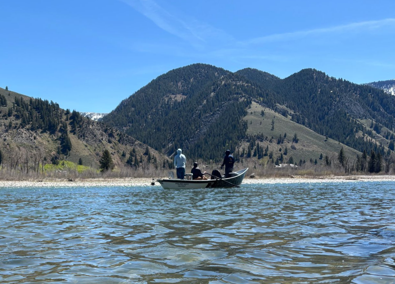Three people on a small boat on a lake with forested mountains under a clear blue sky.