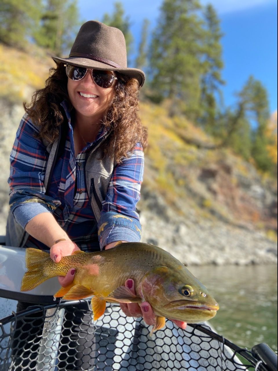 Person holding a large golden trout on a boat by a river, wearing a hat and sunglasses.