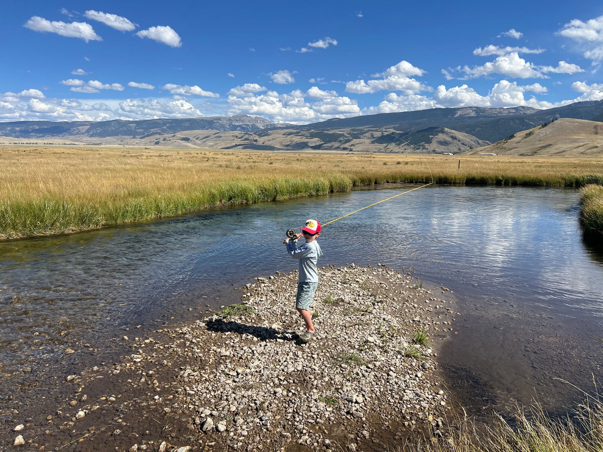 Child fishing in a clear creek beside a grassy plain under a blue sky with mountains